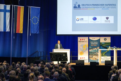 Auf dem Bild sind links drei Flaggen zu sehen: die deutsche, die finnische und die Flagge der EU. Im rechten oberen Quadranten ist ein Slide des Vortrags zu sehen, den der Mann am Rednerpult hält.